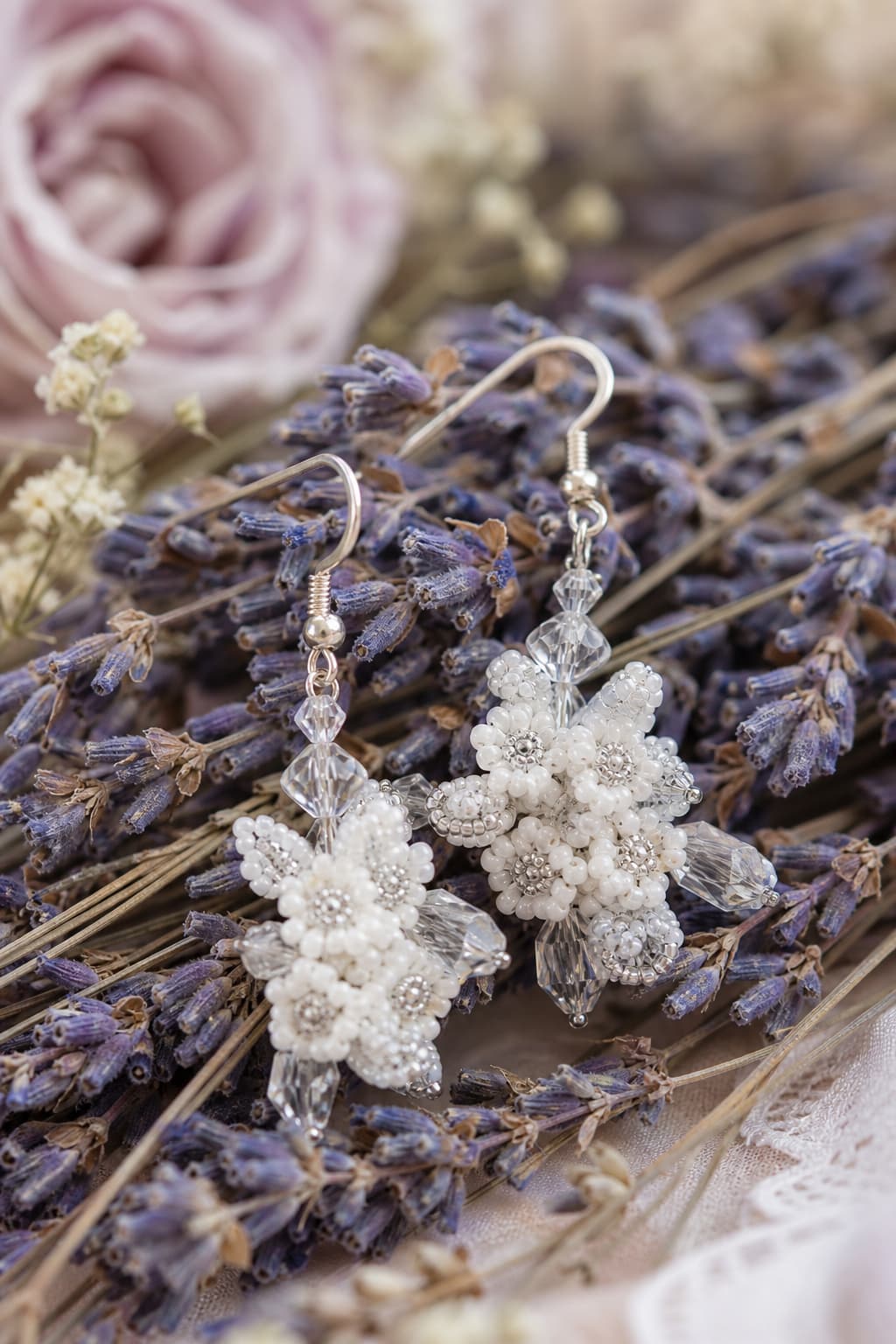 Boucles d’oreilles en forme de fleurs composées de perles de rocaille blanches et de cristaux transparents disposées sur un lit de lavande séchée avec un flou d’arrière-plan doux.