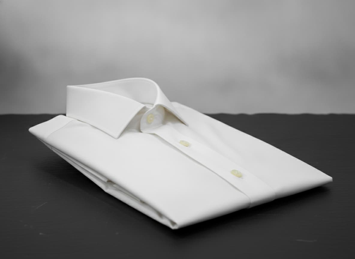 Chemise homme ivoire à col cassé, soigneusement pliée sur une surface ardoise foncée, mettant en valeur la texture du polycoton sous un éclairage élégant.
