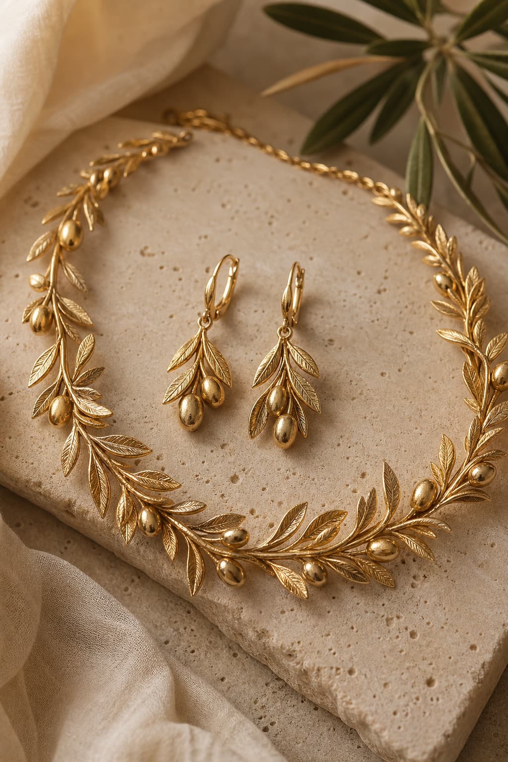 Gros plan d’une parure de mariage dorée composée d’un collier couronne et de boucles d’oreilles en forme de feuilles d’olivier, posée sur une pierre en travertin beige, mettant en valeur les détails métalliques et les ombres douces.