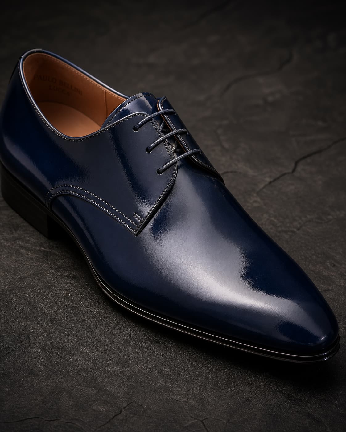 Chaussure de mariage homme en cuir bleu foncé, gros plan incliné sur le bout et les lacets, reflets brillants, éclairage chaleureux sur fond sombre texturé.