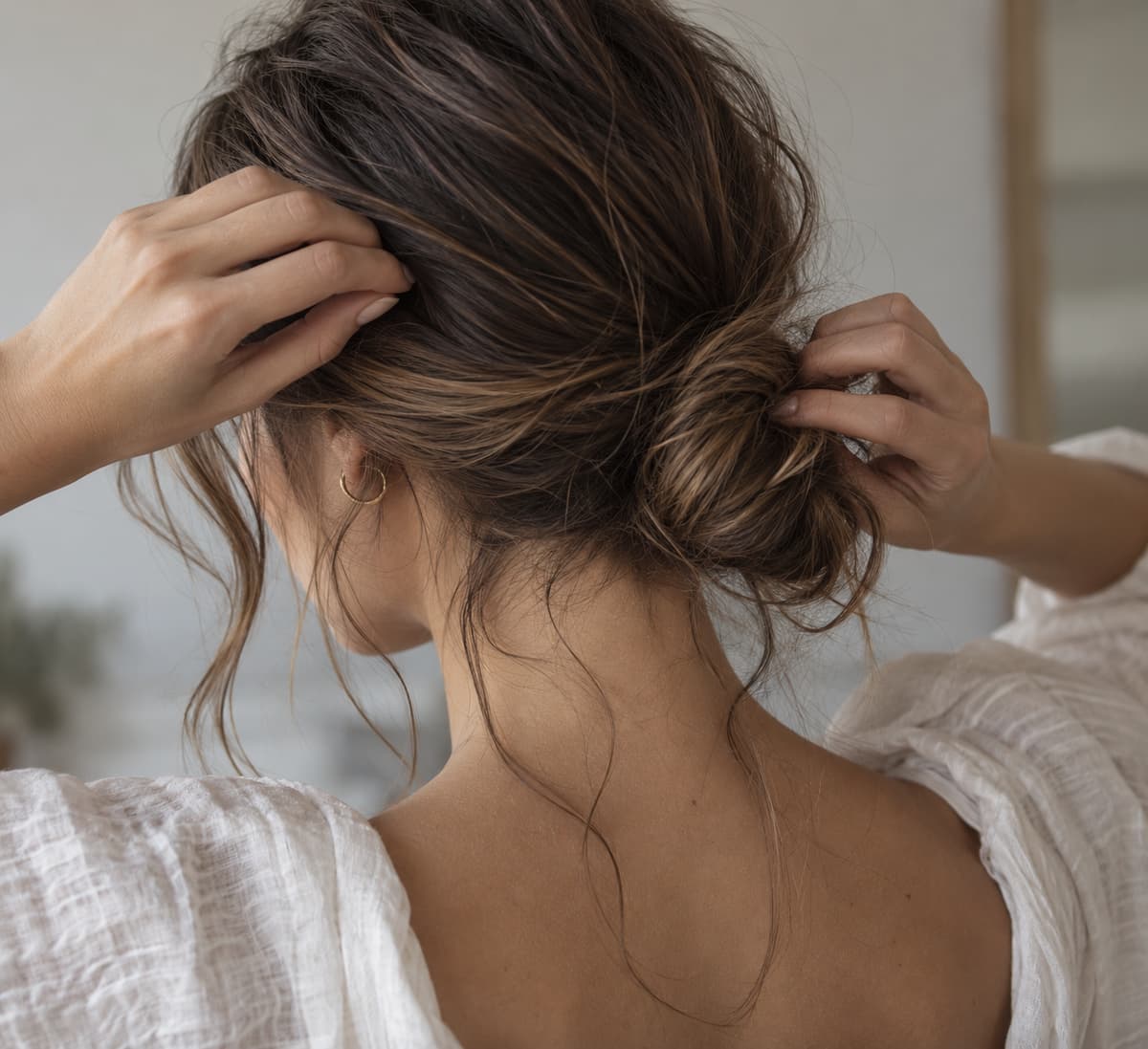 Mains ajustant un chignon bas flou sur la nuque, avec des mèches souples laissées libres et un volume non tiré.
